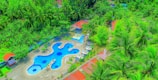 An aerial view of a resort-like area featuring a uniquely shaped blue swimming pool surrounded by lush green vegetation and palm trees. Red-roofed buildings and a series of poolside umbrellas are visible, enhancing the tropical ambiance.
