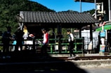 Several children are playing near a small wooden structure with a tiled roof, next to a railing and train tracks. Lush green hills are visible in the background, and colorful signs and posters add vibrancy to the scene.