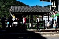Several children are playing near a small wooden structure with a tiled roof, next to a railing and train tracks. Lush green hills are visible in the background, and colorful signs and posters add vibrancy to the scene.