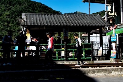 Several children are playing near a small wooden structure with a tiled roof, next to a railing and train tracks. Lush green hills are visible in the background, and colorful signs and posters add vibrancy to the scene.