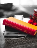 A hardcover book with a red and yellow cover is placed on top of a couple of notebooks on a table. There's a blurred background with a candle and red-and-black patterned object nearby.