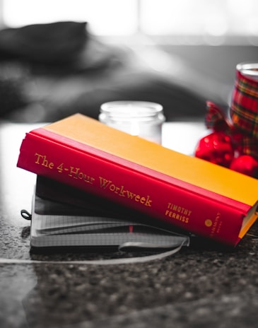 A hardcover book with a red and yellow cover is placed on top of a couple of notebooks on a table. There's a blurred background with a candle and red-and-black patterned object nearby.