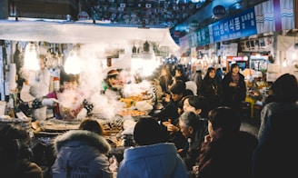 A bustling street food stall is surrounded by a crowd of people. The vendor is busy preparing and serving a variety of dishes. Steam and smoke rise from the hot food, creating a lively and warm atmosphere. The setting appears to be a market with numerous stalls and signage in the background.