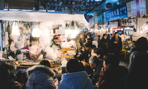 A bustling street food stall is surrounded by a crowd of people. The vendor is busy preparing and serving a variety of dishes. Steam and smoke rise from the hot food, creating a lively and warm atmosphere. The setting appears to be a market with numerous stalls and signage in the background.