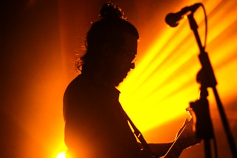 A silhouette of a person playing a guitar is set against a vibrant orange and yellow light. The scene includes a visible microphone stand in the foreground, suggesting a musical performance or concert atmosphere with dramatic lighting.