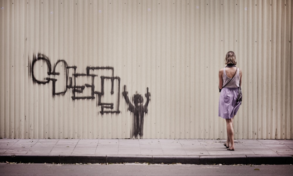 白いタンクトップと紫色のスカートを着た女性が、落書きで描かれた壁に面して立っている