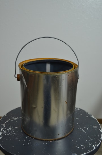 A metallic paint can with rust stains around the rim sits on a black, slightly scratched surface. The can is open with its handle upright.