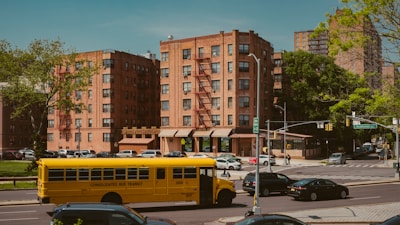A yellow school bus drives down a street lined with parked cars beside a series of brick apartment buildings. The architecture features fire escapes and there are trees and a traffic light at the intersection.