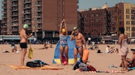 A beach scene featuring people in swimsuits with swimming caps and gear. In the background, there are tall apartment buildings and people relaxing on the sand.