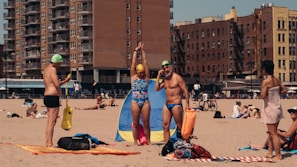 A beach scene featuring people in swimsuits with swimming caps and gear. In the background, there are tall apartment buildings and people relaxing on the sand.