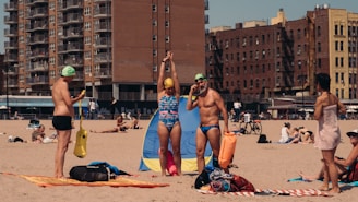 A beach scene featuring people in swimsuits with swimming caps and gear. In the background, there are tall apartment buildings and people relaxing on the sand.
