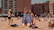 A beach scene featuring people in swimsuits with swimming caps and gear. In the background, there are tall apartment buildings and people relaxing on the sand.