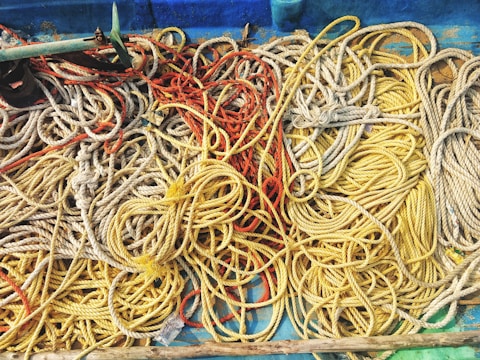 A tangled collection of ropes in various colors, including yellow, red, and beige, lie strewn across a blue surface. The ropes appear intertwined and haphazardly arranged, suggesting the cluttered setting of a boat or dock area.