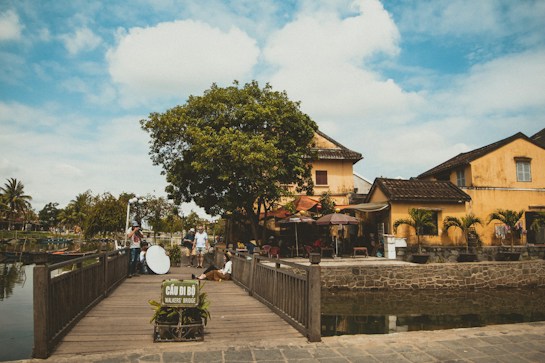 A scenic view of a small, wooden pedestrian bridge leading to a quaint area with yellow, traditional-looking buildings. A vibrant tree provides shade, and people are using the bridge. A sign on the bridge reads 'Cầu đi bộ - Walkers' Bridge.' The sky is partially cloudy, contributing to a serene atmosphere.