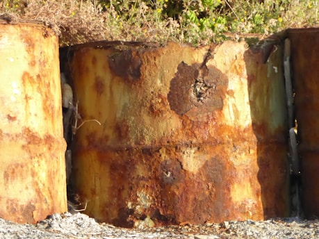 Three large, rusted metal barrels appear partially buried in the ground. The surface of the barrels is heavily corroded, displaying a mix of orange and brown hues. Overgrown vegetation surrounds the barrels, indicating they have been in place for a long time.