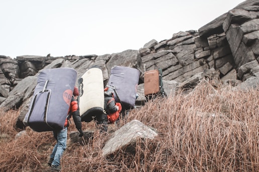 Several individuals, equipped with large crash pads and backpacks, are climbing a rocky hill. The terrain is rugged, with dry grass and prominent rock formations. The group appears to be on a hiking or bouldering expedition.