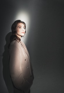 A person is standing in a spotlight with a dark background, wearing a light-colored suit. The lighting creates a dramatic shadow on the wall, and the person looks contemplative.