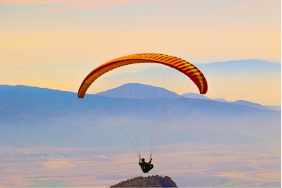 A person is paragliding against a backdrop of distant mountains and a hazy sky. The paraglider canopy is multicolored with shades of orange and yellow, and the person is sitting in a harness beneath it. The scene conveys a sense of freedom and adventure.