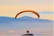 A person is paragliding against a backdrop of distant mountains and a hazy sky. The paraglider canopy is multicolored with shades of orange and yellow, and the person is sitting in a harness beneath it. The scene conveys a sense of freedom and adventure.