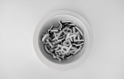 A white bowl filled with black and white alphabet letters is placed on a plain white surface. The letters appear to be made of plastic and are randomly arranged within the bowl.