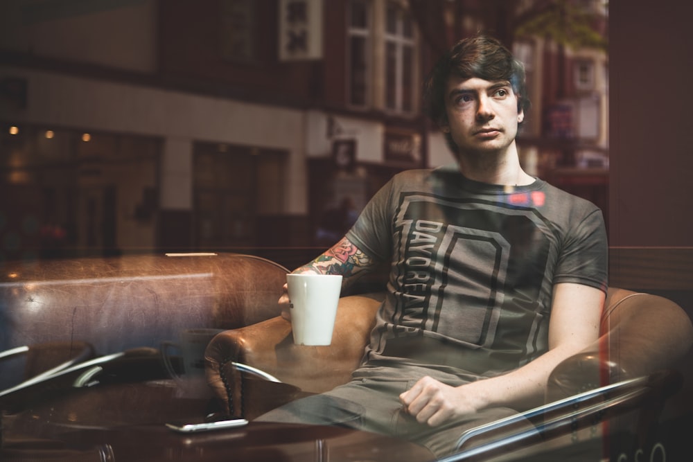 man sitting on beige armchair while holding white ceramic mug