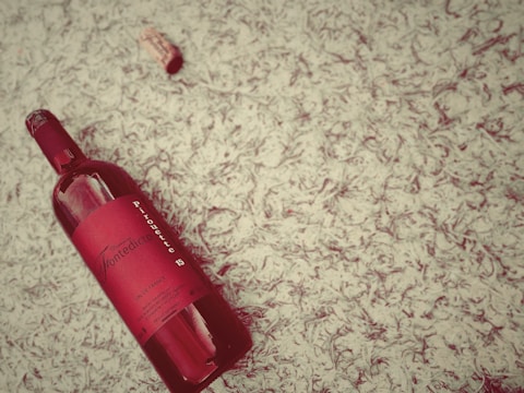 A red wine bottle with a detailed label is lying on a textured surface, accompanied by a cork nearby. The label reads 'Domaine Fontedicto' and 'Pirouette' among other information, and both the bottle and cork are positioned at an angle. The background features a cream-colored, leafy pattern.