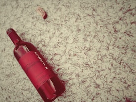 A red wine bottle with a detailed label is lying on a textured surface, accompanied by a cork nearby. The label reads 'Domaine Fontedicto' and 'Pirouette' among other information, and both the bottle and cork are positioned at an angle. The background features a cream-colored, leafy pattern.
