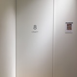 A plain light-colored wall features a door labeled with the number 8 and the word 'CRAFT' in bold black letters. To the right, a bulletin or flyer is affixed to the wall with a small colorful image and text.