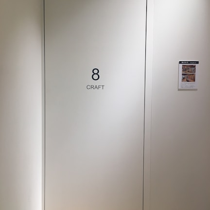 A plain light-colored wall features a door labeled with the number 8 and the word 'CRAFT' in bold black letters. To the right, a bulletin or flyer is affixed to the wall with a small colorful image and text.