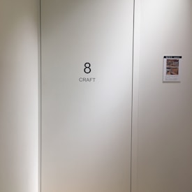 A plain light-colored wall features a door labeled with the number 8 and the word 'CRAFT' in bold black letters. To the right, a bulletin or flyer is affixed to the wall with a small colorful image and text.
