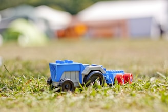 A plastic toy vehicle, resembling a tractor or construction equipment, made from colorful building blocks is placed on a grassy surface. The vehicle is primarily blue with accents of gray and red, and has large black wheels. In the blurred background, tents or structures can be seen, suggesting an outdoor setting.