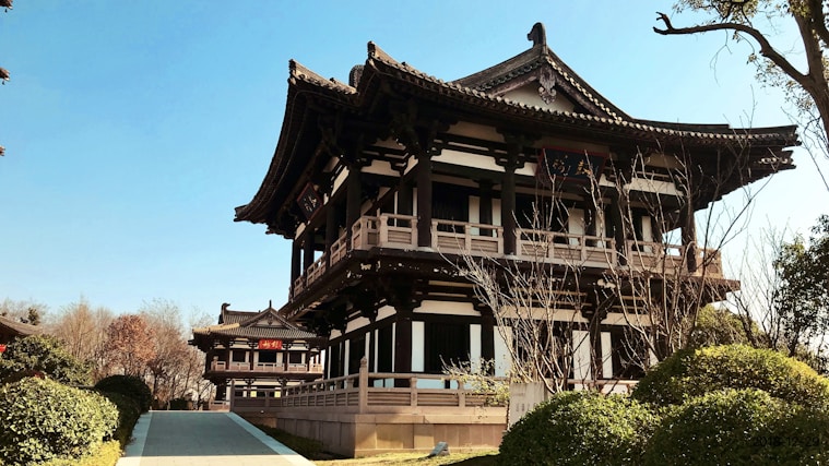 An intricately designed traditional building with multiple eaves and ornate carvings is situated in a serene environment. Shrubs and trees surround it, and there's a pathway leading up to the structure. The sky is clear and blue, emphasizing the tranquil ambiance.