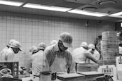 A group of people wearing uniforms and caps are working together in a kitchen setting. They are focused on preparing food at a counter, with steaming baskets and various kitchen items around them. The environment appears organized and busy, with tiled walls and overhead lights.