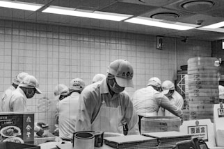 A group of people wearing uniforms and caps are working together in a kitchen setting. They are focused on preparing food at a counter, with steaming baskets and various kitchen items around them. The environment appears organized and busy, with tiled walls and overhead lights.