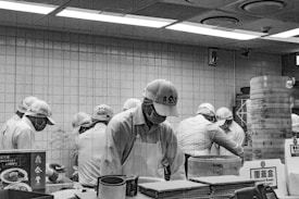 A group of people wearing uniforms and caps are working together in a kitchen setting. They are focused on preparing food at a counter, with steaming baskets and various kitchen items around them. The environment appears organized and busy, with tiled walls and overhead lights.