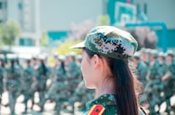 A group of people in military camouflage uniforms stand in formation, one in the foreground with long hair visible from behind. The scene appears to be a training exercise or ceremonial lineup with a blurred background suggesting outdoor setting near buildings and trees.