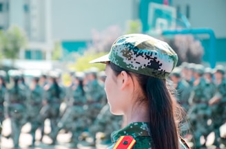 A group of people in military camouflage uniforms stand in formation, one in the foreground with long hair visible from behind. The scene appears to be a training exercise or ceremonial lineup with a blurred background suggesting outdoor setting near buildings and trees.