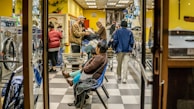 A busy laundromat with several people engaged in doing laundry. Some are standing by the machines, others are walking with laundry baskets. A person is seated on a bench, appearing to wait. The interior is bright with yellow walls and gray checkered flooring. Various items of clothing and laundry baskets are visible throughout the space.
