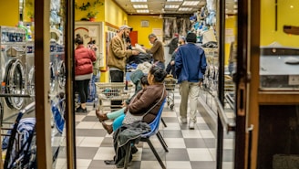 A busy laundromat with several people engaged in doing laundry. Some are standing by the machines, others are walking with laundry baskets. A person is seated on a bench, appearing to wait. The interior is bright with yellow walls and gray checkered flooring. Various items of clothing and laundry baskets are visible throughout the space.