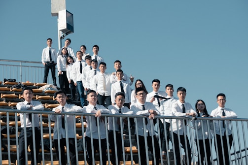 A group of students in white shirts and black ties stands on outdoor bleachers under a clear blue sky. They are posing for a group photo, with some resting their arms on a metal railing in the foreground. The atmosphere appears to be a mix of playful and formal poses.