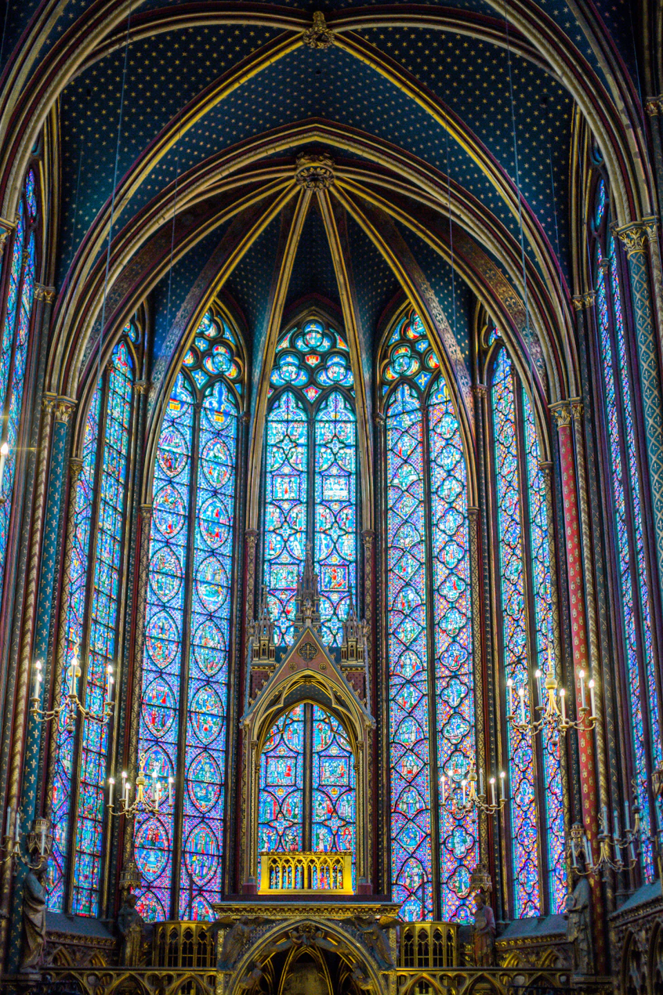 The image depicts a large, ornate cathedral with a high ceiling and multiple arched windows adorned with stained glass panels. The windows are predominantly blue and green hues, creating a vibrant contrast against the stone walls. At the center of the cathedral is a large, ornate altar, which serves as the focal point for religious ceremonies within the space.

The cathedral features several statues scattered throughout its interior, adding to the overall grandeur of the scene. The perspective of the image is from below, looking up at the towering ceiling and intricate details of the stained glass windows and stone walls.