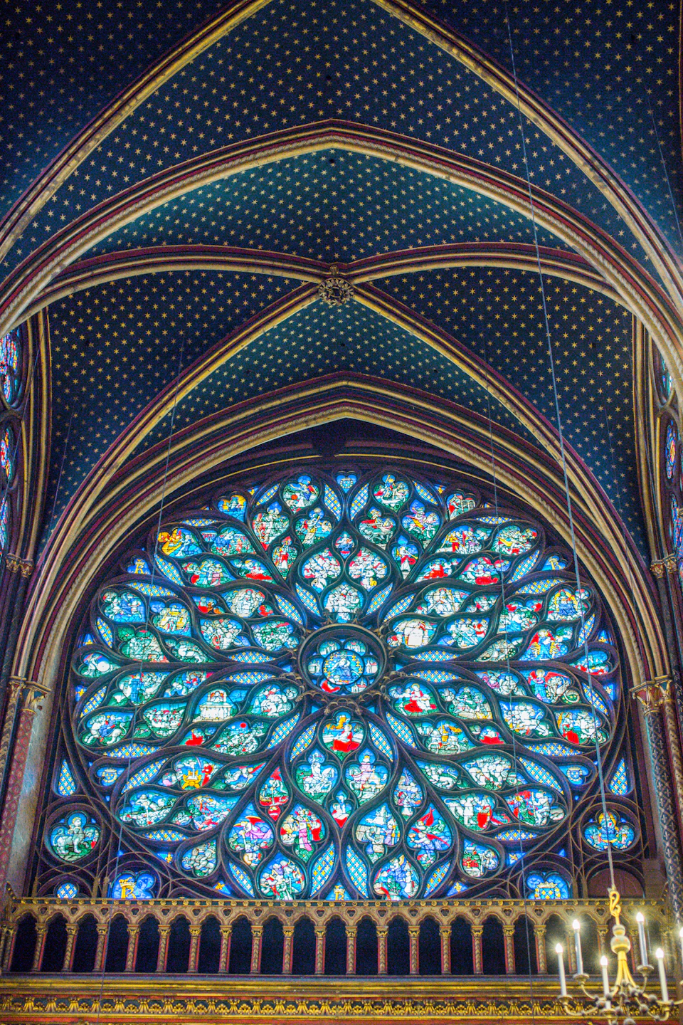 The image shows a large, ornate stained glass window situated within an architectural space with a high ceiling and pointed arches. The window is a riot of colors, featuring intricate designs that are likely to bewitch viewers with their beauty. The light filtering through the stained glass creates a kaleidoscope of hues, casting a warm glow throughout the room.

The perspective of the image is from below, looking up at the window, which enhances its grandeur and magnifies its details. The high ceiling adds an air of majesty to the scene, making it feel like one is standing in the heart of a cathedral or a palace.