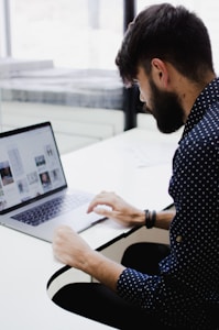 A person with a beard is sitting at a white desk, focused on a laptop screen displaying multiple images and text. The individual is wearing a dark shirt with white polka dots. The environment appears to be a modern office or workspace with bright natural light coming through large windows.