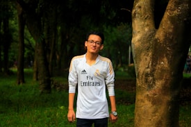 A person wearing a white Adidas football jersey with the Real Madrid emblem stands next to a large tree. The background features a lush, green forest with diffused light creating a serene atmosphere.
