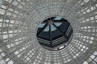 A circular, intricate glass ceiling design with a central opening, featuring a repetitive geometric pattern of small, textured glass squares. The design creates an illusion of depth and symmetry, with reflections playing across the surface.