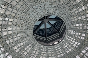 A circular, intricate glass ceiling design with a central opening, featuring a repetitive geometric pattern of small, textured glass squares. The design creates an illusion of depth and symmetry, with reflections playing across the surface.