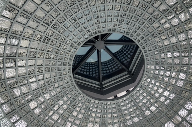 A circular, intricate glass ceiling design with a central opening, featuring a repetitive geometric pattern of small, textured glass squares. The design creates an illusion of depth and symmetry, with reflections playing across the surface.