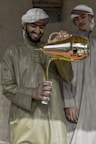 Two men in traditional attire are engaged in pouring a beverage from a golden teapot into a decorated cup. The man holding the teapot is smiling, suggesting a friendly or hospitable atmosphere. They are standing against a rustic background.