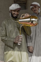 Two men in traditional attire are engaged in pouring a beverage from a golden teapot into a decorated cup. The man holding the teapot is smiling, suggesting a friendly or hospitable atmosphere. They are standing against a rustic background.