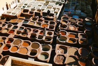A traditional tannery with numerous circular pits filled with various colored dyes and tanned hides. Workers are seen moving between the pits, tending to the hides, and using long sticks. The scene is bathed in natural light, highlighting the rich and earthy tones of the dyes and leather.